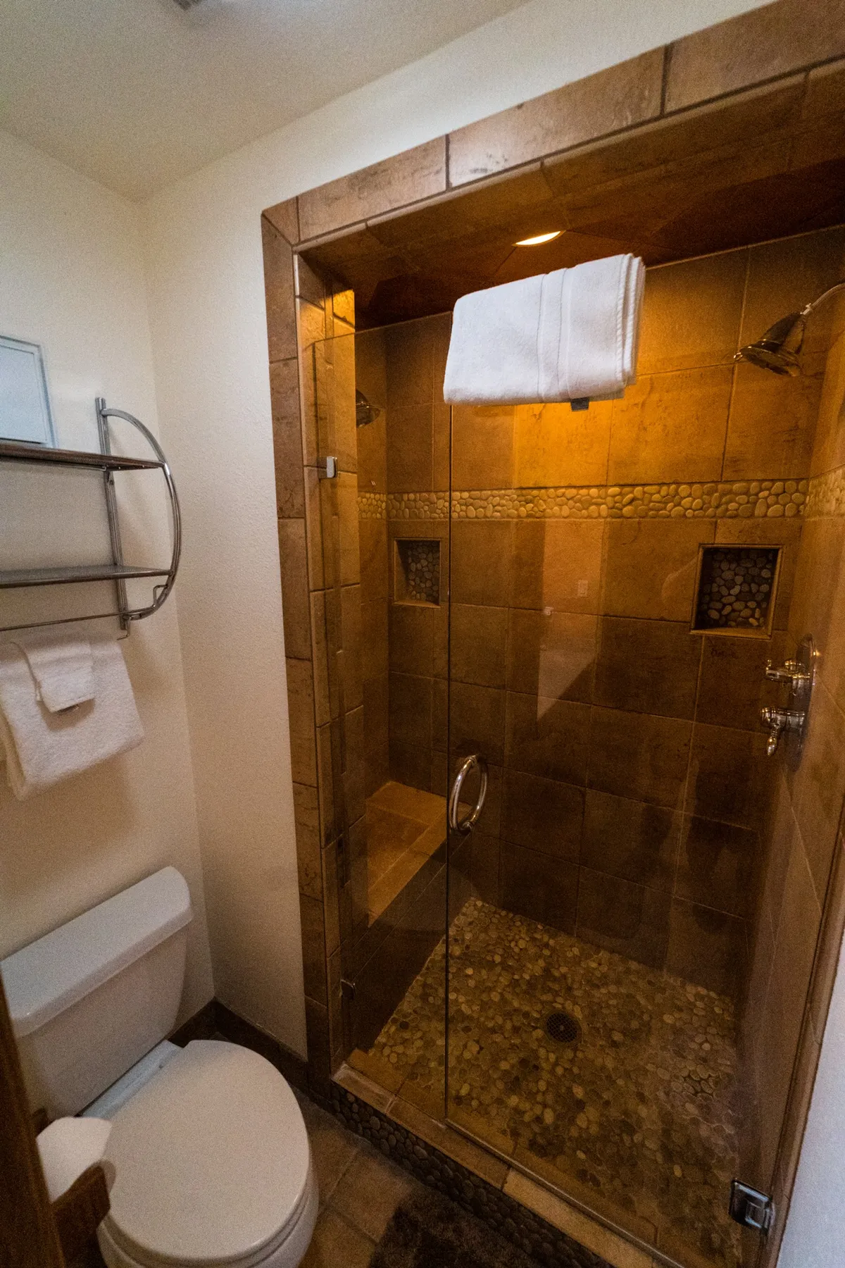 A modern en-suite bathroom featuring a glass-enclosed shower with pebble rock flooring, a towel rack with fluffy towels, and a calm, spa-like atmosphere. The view focuses on the shower area, emphasizing the multi-head shower system and serene design.