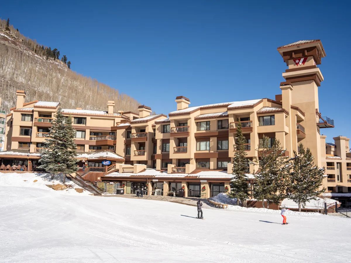 The exterior shot highlights the ski resort building surrounded by snowy slopes and trees. The image conveys an atmosphere of adventure and comfort, with visible balconies that hint at where guests can relax after days on the mountain.