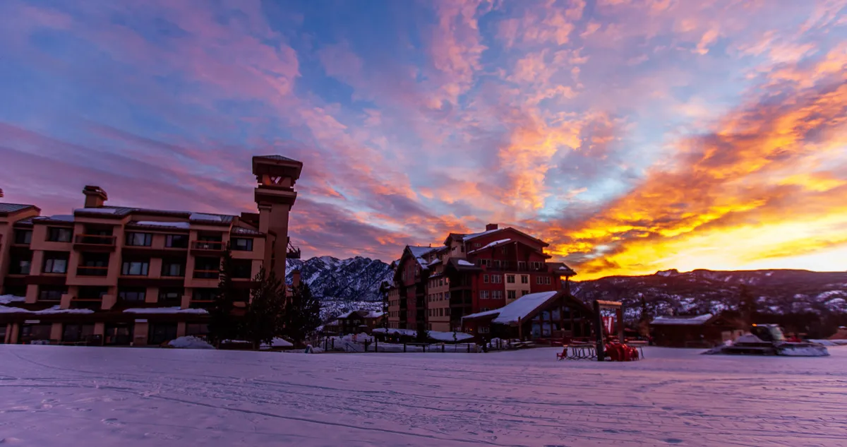 This exterior scene showcases a tranquil, snow-covered landscape with colorful sunset hues illuminating the buildings. The lodge is inviting and there’s a sense of calm as the day winds down, ideal for capturing the mood of relaxation after action-packed activities.