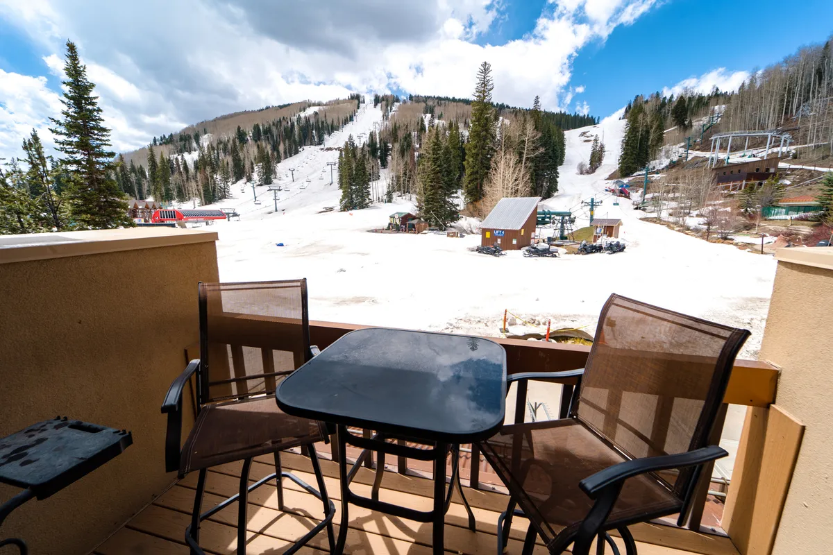 A private balcony with comfortable seating overlooking a snow-covered mountain landscape, chairlifts in the near distance, and skiers passing by. The balcony is ideal for enjoying morning coffee, grilling on the BBQ, or relaxing at sunset while immersed in the alpine setting.