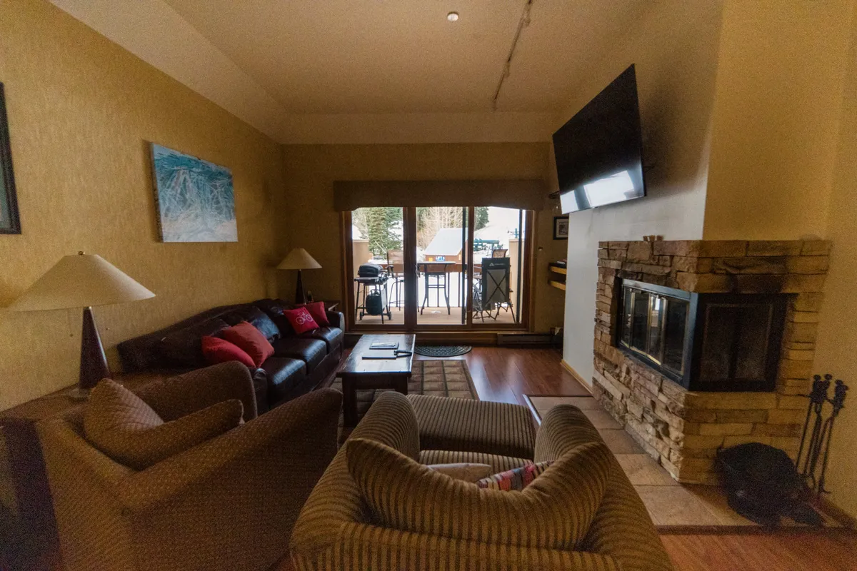 A welcoming living room with a cozy wood-burning fireplace, comfortable sofa seating arranged for conversation or relaxation, and expansive windows that frame the snow-covered slopes and nearby chairlifts. The atmosphere suggests warmth and comfort, with natural light streaming in and a perfect view for après-ski lounging.