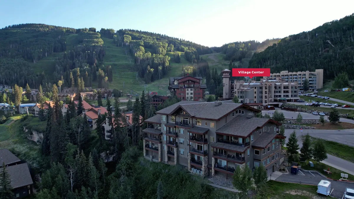 The image highlights the modern Village Center set against a dramatic mountain backdrop, with the 'Village Center' sign, pine trees, and hints of activity. The scene evokes the allure of exclusive resort amenities and a welcoming après-ski environment.