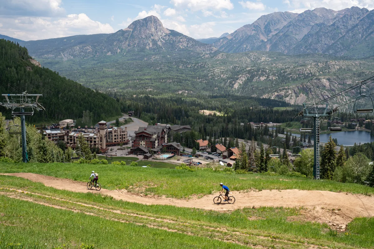 A breathtaking view of the San Juan mountain range with resort buildings nestled among biking trails and a tranquil lake in the foreground. The expansive landscape evokes a sense of adventure and relaxation, showcasing opportunities for mountain biking, hiking, and exploring nature just outside the condo.