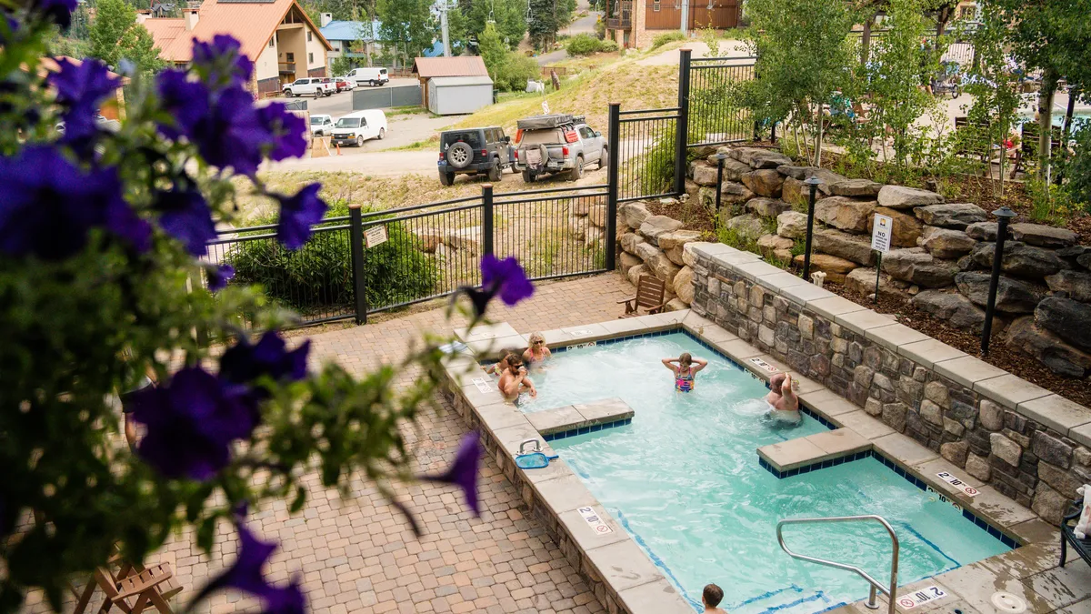 This exterior image showcases a shared hot tub and swimming pool area surrounded by lush foliage, with people enjoying both the pool and the hot tub. The scene feels bright, lively, and communal, reflecting a resort-like atmosphere.