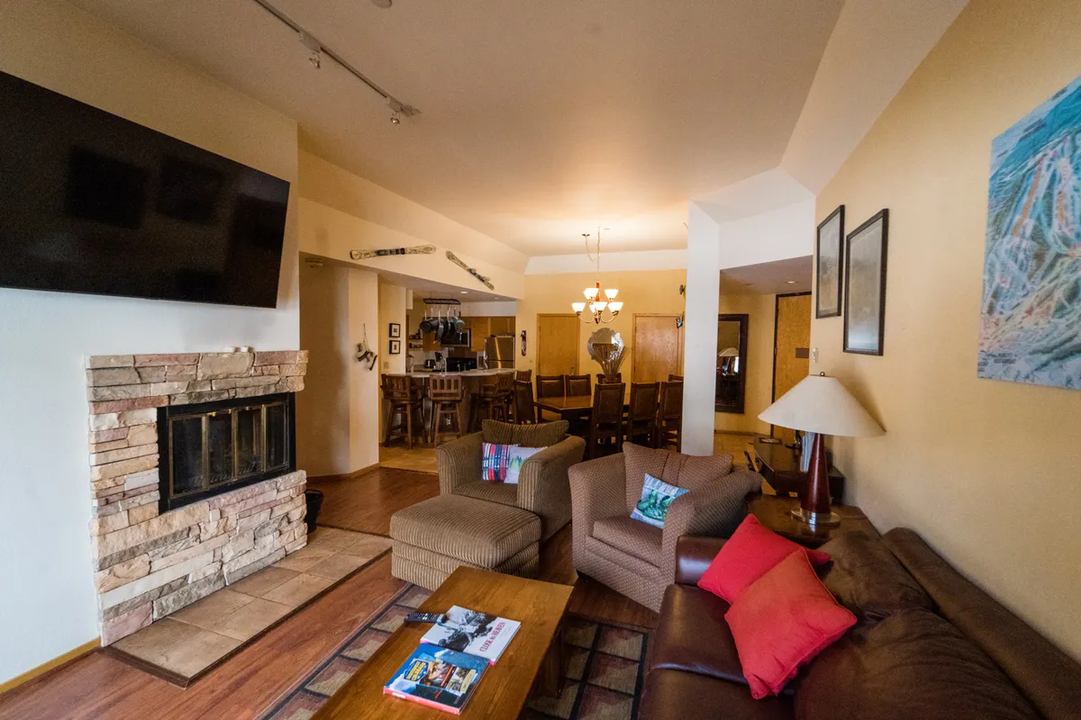 The image showcases the inviting living area of the condo, with plush sofas arranged around a glowing fireplace and a large TV. A stylish dining area is visible nearby, creating a warm space perfect for gathering with friends and family after a day on the slopes. The mood is cozy and sociable, ideal for relaxing, sharing stories, and enjoying special moments together.