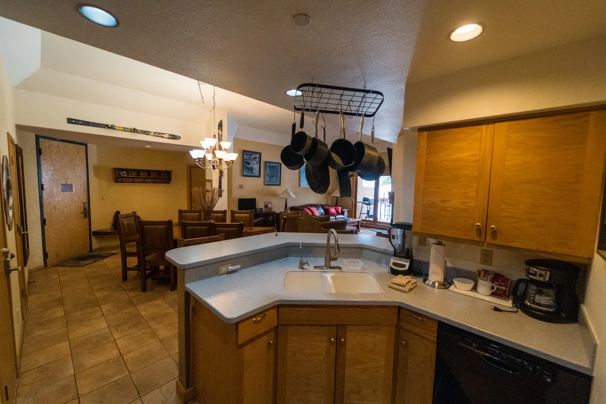 A warm and cozy kitchen scene centered on a spacious island, with hanging pots overhead and clear views to the adjacent dining area and living room. The space is designed for active use, with plenty of room for cooking, dining, and gathering.