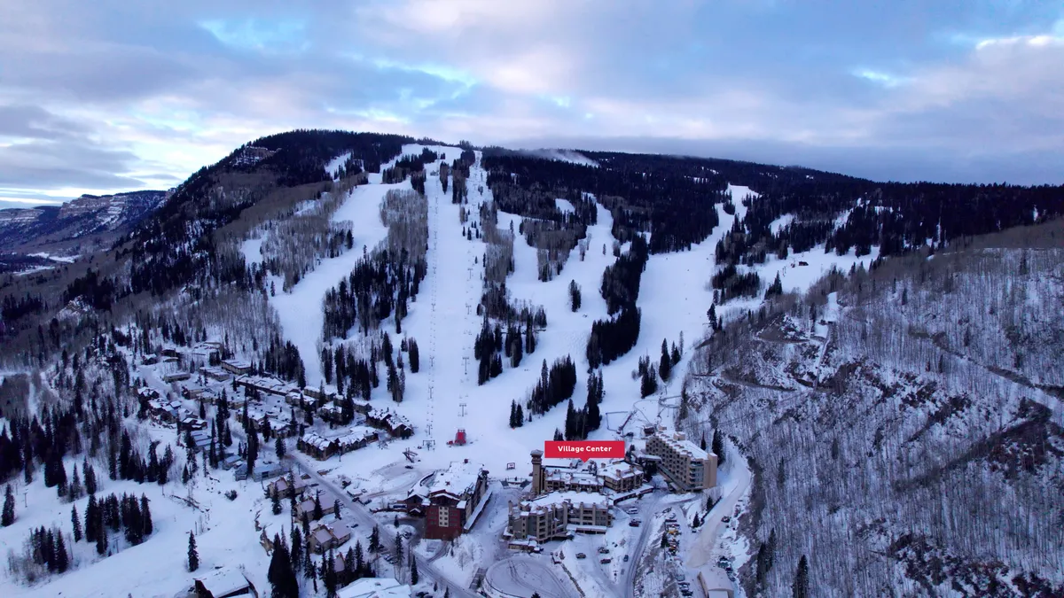 This exterior shot captures the snowy slopes, pine trees, and central location of the Village Center. Skiers and snowboarders can be seen enjoying the terrain, with the village itself offering a cozy hub for après-ski gatherings.