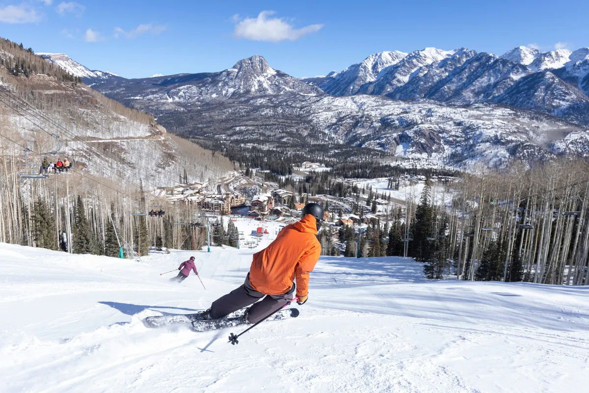 This wide view shows skiers in action on the slopes, snow-covered mountains, and a ski lift ascending in the background. The image encapsulates the thrill and beauty of the resort’s winter sports environment.