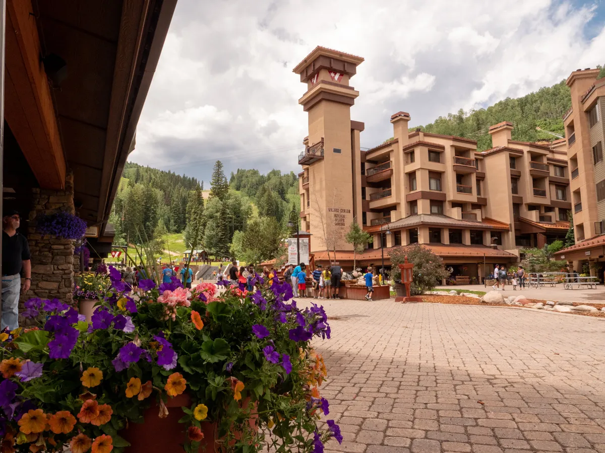 This exterior image features the vibrant Village Center building, complete with a distinctive clock tower, surrounded by lush flower beds and people enjoying the lively communal spaces. The scene communicates an inviting, family-friendly atmosphere ideal for making memories together.