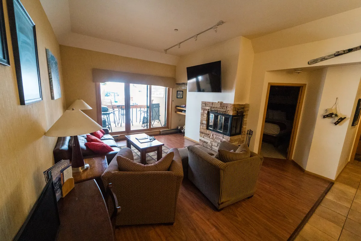 A cozy mountain-style living room featuring a stone fireplace, large windows that flood the space with natural light, comfortable seating, a TV, and a coffee table. The windows offer scenic mountain views, and the space is ideal for relaxing after outdoor adventures or enjoying a sunset from the balcony.