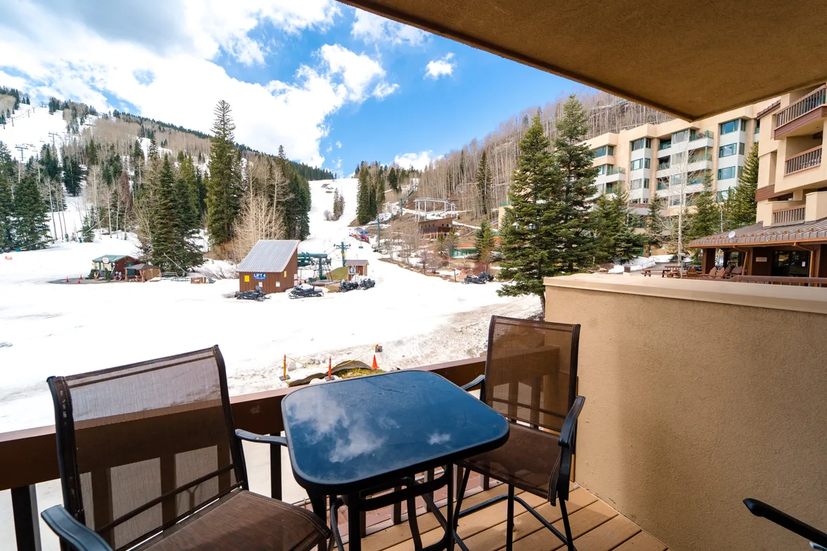 This image captures an inviting outdoor seating area overlooking snow-covered slopes, with a backdrop of mountains and trees. The building facade showcases the close proximity to the ski runs, and the scene suggests both relaxation and easy access to winter sports.