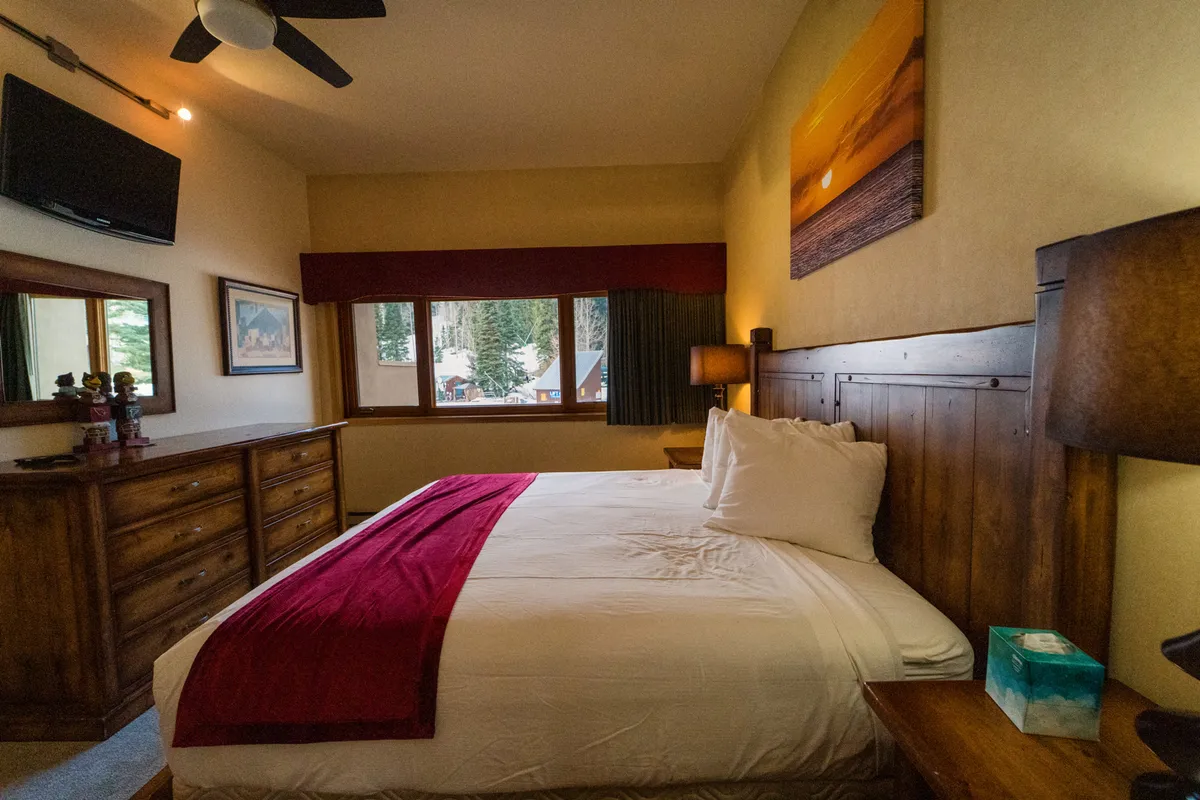 A serene king bedroom featuring a plush bed with a red accent blanket, large windows offering scenic views, a wooden dresser, and tasteful wall art. The room is well-lit and exudes a calming, comfortable atmosphere, with blackout blinds partially visible for privacy and rest.