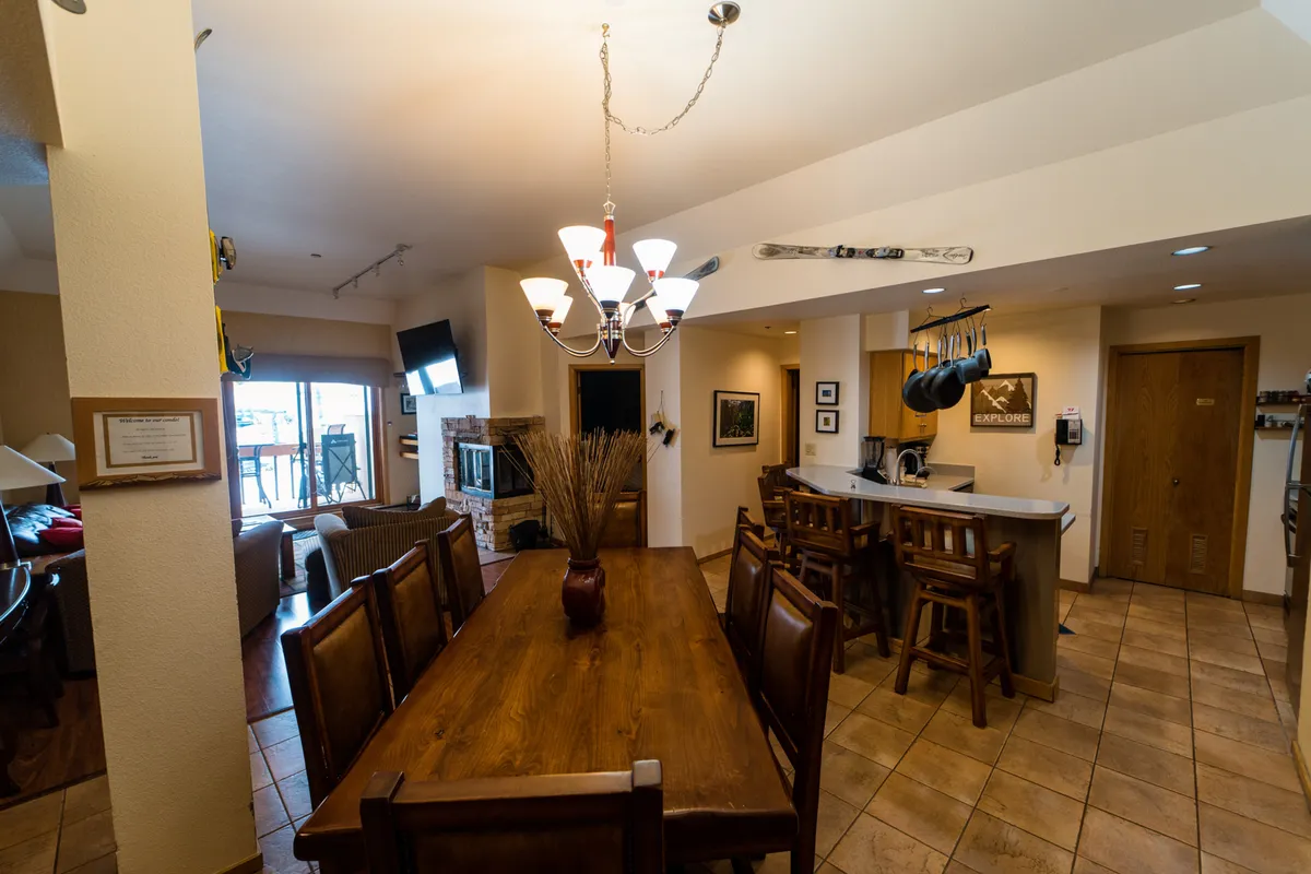 A spacious dining area featuring a large wooden dining table, an elegant chandelier overhead, a view of the adjacent kitchen bar, and a glimpse of the fireplace. The scene feels perfect for gathering friends or family, sharing meals, and enjoying the warmth of the space after a day on the slopes.